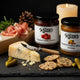 Mrs. Ruth's Roasted Tomato Chutney and Roasted Pineapple Habanero Chutney jars standing upright on a dark slate surface surrounded by a kraft gift box, aged manchego, seeded crackers, cornichons, prosciutto, and rosemary sprigs in warm candlelight