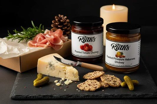 Mrs. Ruth's Roasted Tomato Chutney and Roasted Pineapple Habanero Chutney jars standing upright on a dark slate surface surrounded by a kraft gift box, aged manchego, seeded crackers, cornichons, prosciutto, and rosemary sprigs in warm candlelight