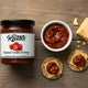 Flat lay of Mrs. Ruth's Roasted Tomato Chutney jar next to a ramekin of chunky red chutney, aged cheddar wedge, and seeded crackers topped with cheddar and chutney on a rustic wooden surface