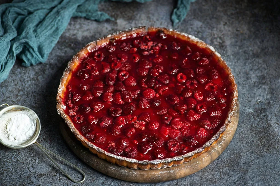 A whole Mrs. Ruth's Raspberry Tart with a golden buttery crust, topped with fresh raspberries baked in a glossy Special Reserve Raspberry Jam glaze, on a table with a powdered sugar sifter alongside