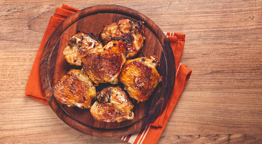 Pieces of baked chicken glazed with Mrs. Ruth's Balsamic Strawberry Jam arranged on a round rustic wooden serving tray, garnished with fresh sliced strawberries and herbs, with the Balsamic Strawberry Jam jar in the background