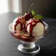 Two scoops of vanilla ice cream in a glass sundae dish topped with warm Mrs. Ruth's Belgian Chocolate Cherry Jam sauce, toasted almonds, flaky sea salt, and a single fresh cherry, on a dark slate surface
