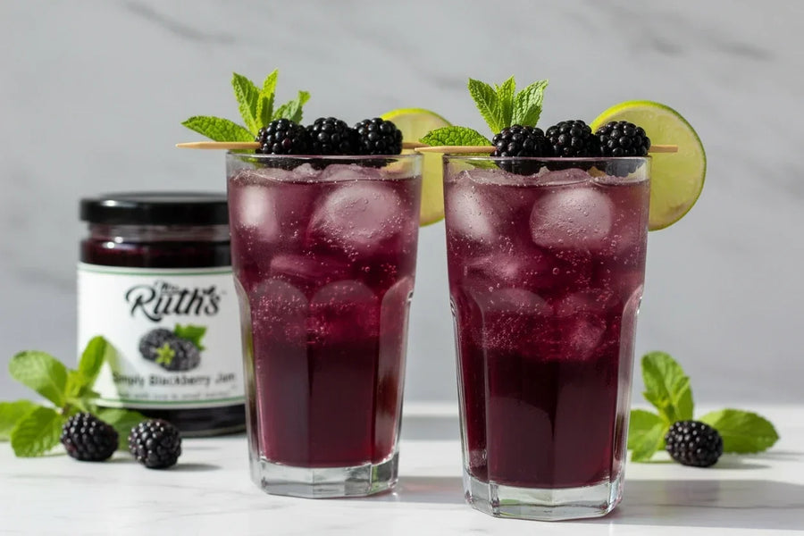Two tall glasses filled with a jewel-toned purple blackberry mojito mocktail over ice, garnished with fresh blackberries, mint sprigs, and a lime wedge, with a Mrs. Ruth's Simply Blackberry Jam jar softly blurred in the background