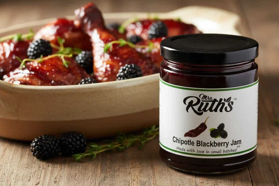Mrs. Ruth's Chipotle Blackberry Jam jar in sharp focus in the foreground with a rustic casserole dish of chipotle blackberry glazed chicken thighs softly blurred behind it, fresh blackberries and thyme scattered on a wooden surface