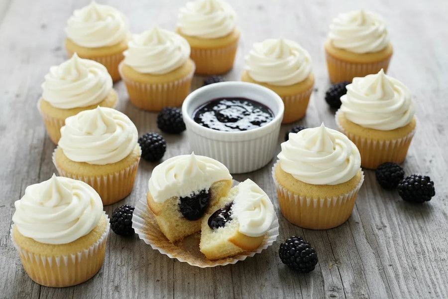 Golden vanilla cupcakes with cream cheese frosting scattered on a rustic wooden surface, one cut open to reveal a glossy blackberry jam filling, with a small bowl of Mrs. Ruth's Simply Blackberry Jam and fresh blackberries alongside