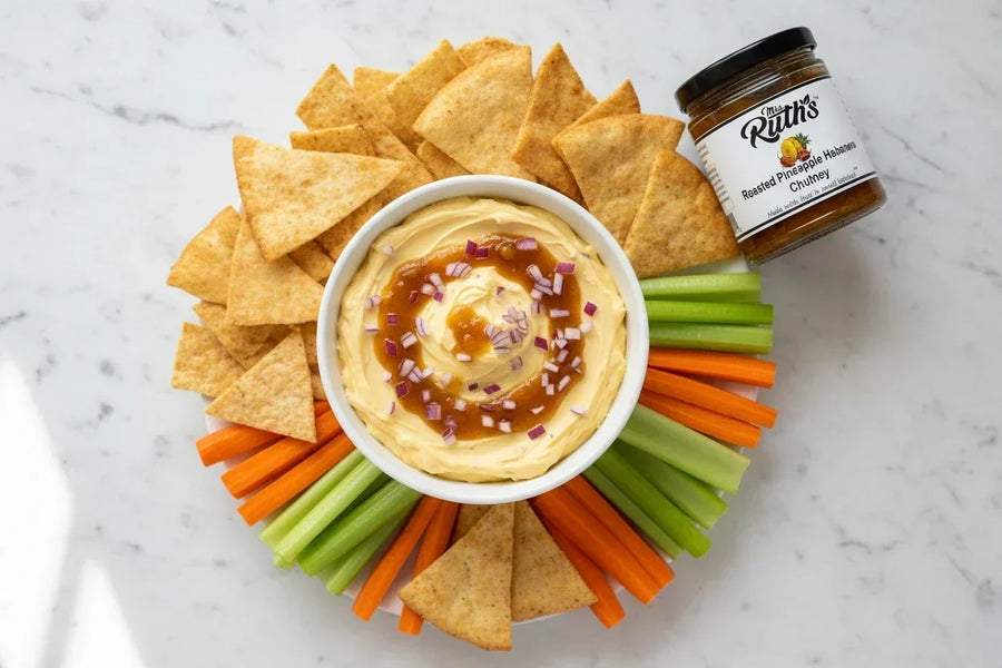 Overhead view of a creamy pineapple habanero cream cheese dip in a white bowl, drizzled with Mrs. Ruth's Roasted Pineapple Habanero Chutney, surrounded by crackers, pita chips, celery, and carrots on a white marble surface